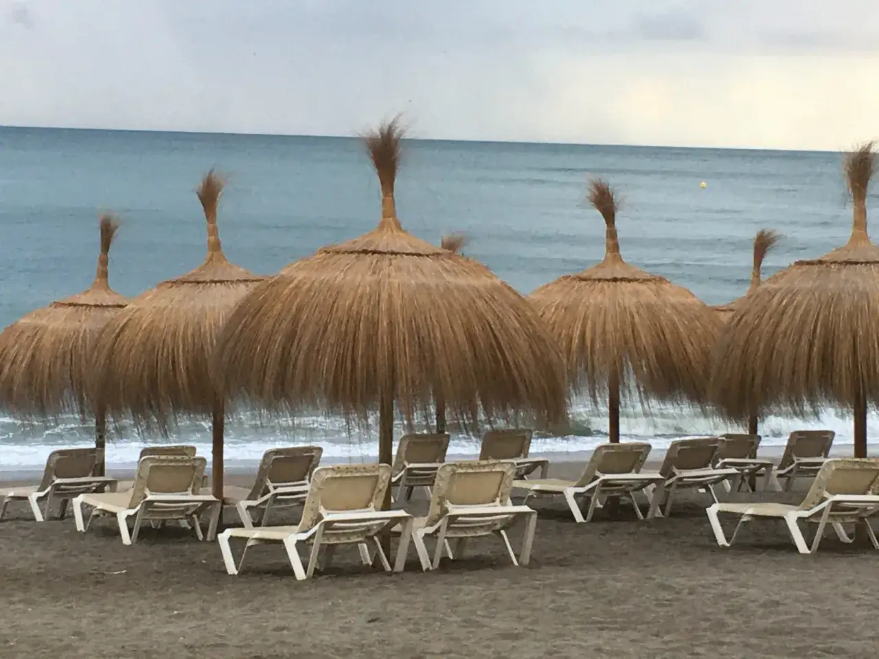 Plaża w Maladze we wrześniu, puste leżaki pod słomianymi parasolami, morze i zachmurzone niebo.