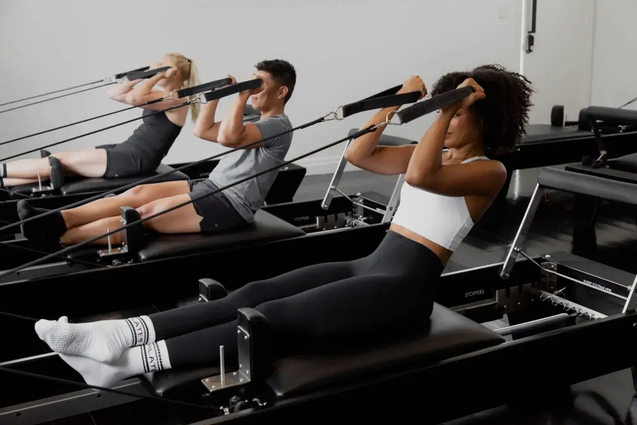 Tre personer tr&auml;nar reformer pilates. De ligger p&aring; rygg och drar i handtag med rep.