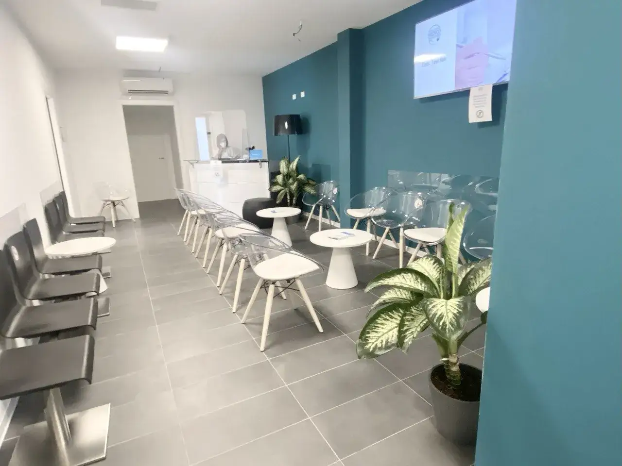 Sala d'attesa moderna, con sedie trasparenti e scure, tavolini, piante e un bancone reception. Si percepisce la differenza tra ambulatorio e poliambulatorio.