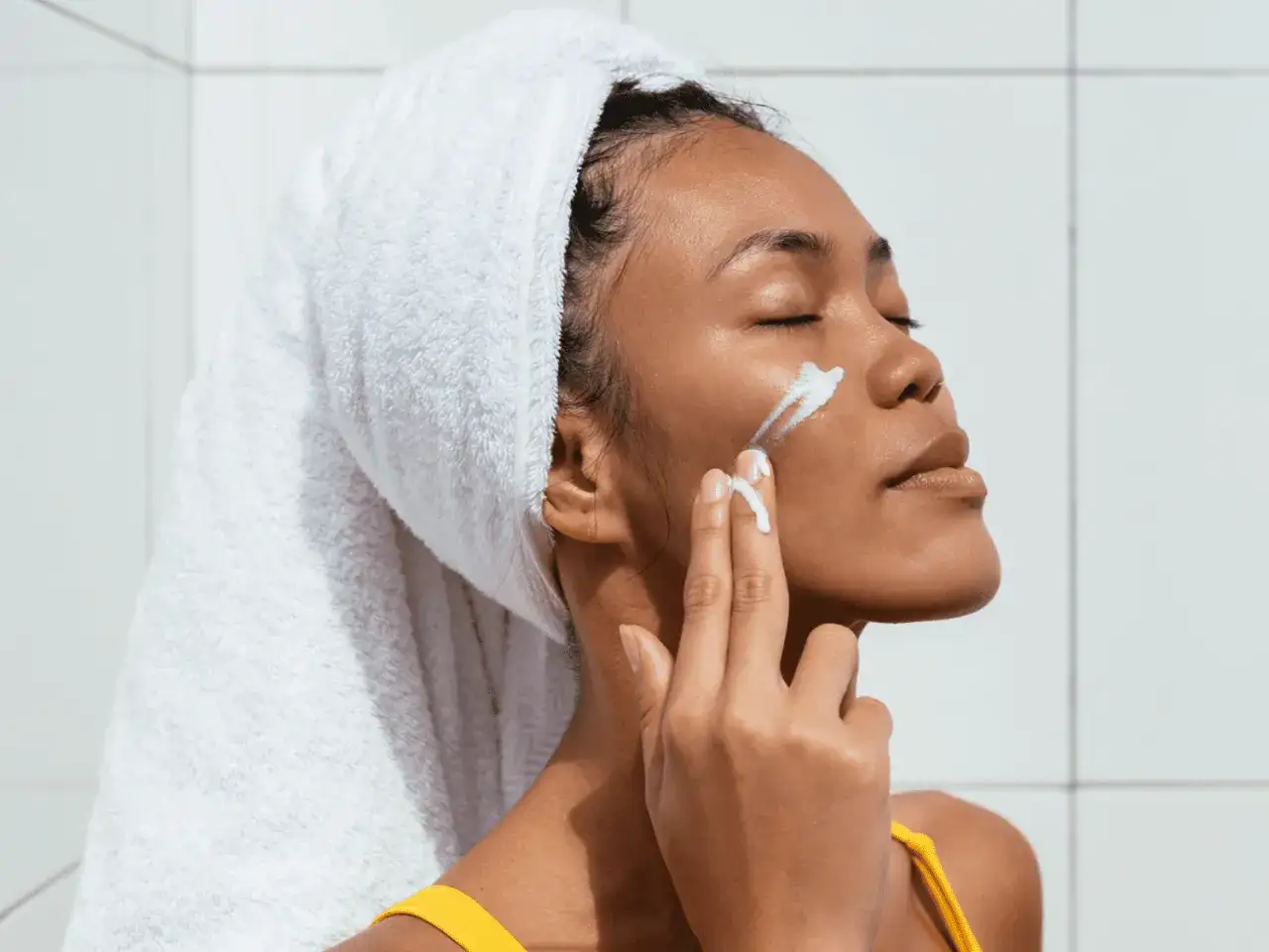 Mujer con toalla en la cabeza se aplica crema en la cara. Piensa en qué pasa si no te pones crema solar.