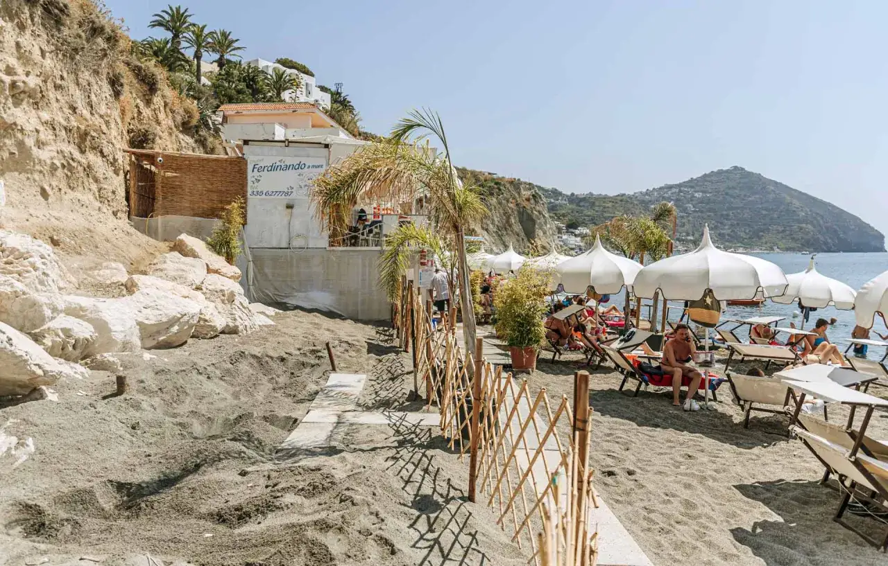 Spiaggia dei Maronti, lidi con ombrelloni bianchi e lettini, gente che si rilassa al sole. Un ristorante con insegna