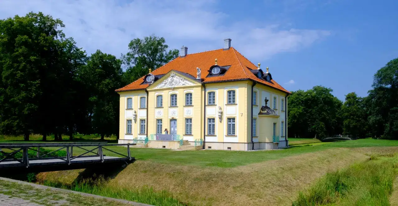 Ż&oacute;łty pałac w Choroszczy z pomarańczowym dachem, otoczony zielenią i fosą. Drewniany mostek prowadzi do posiadłości.