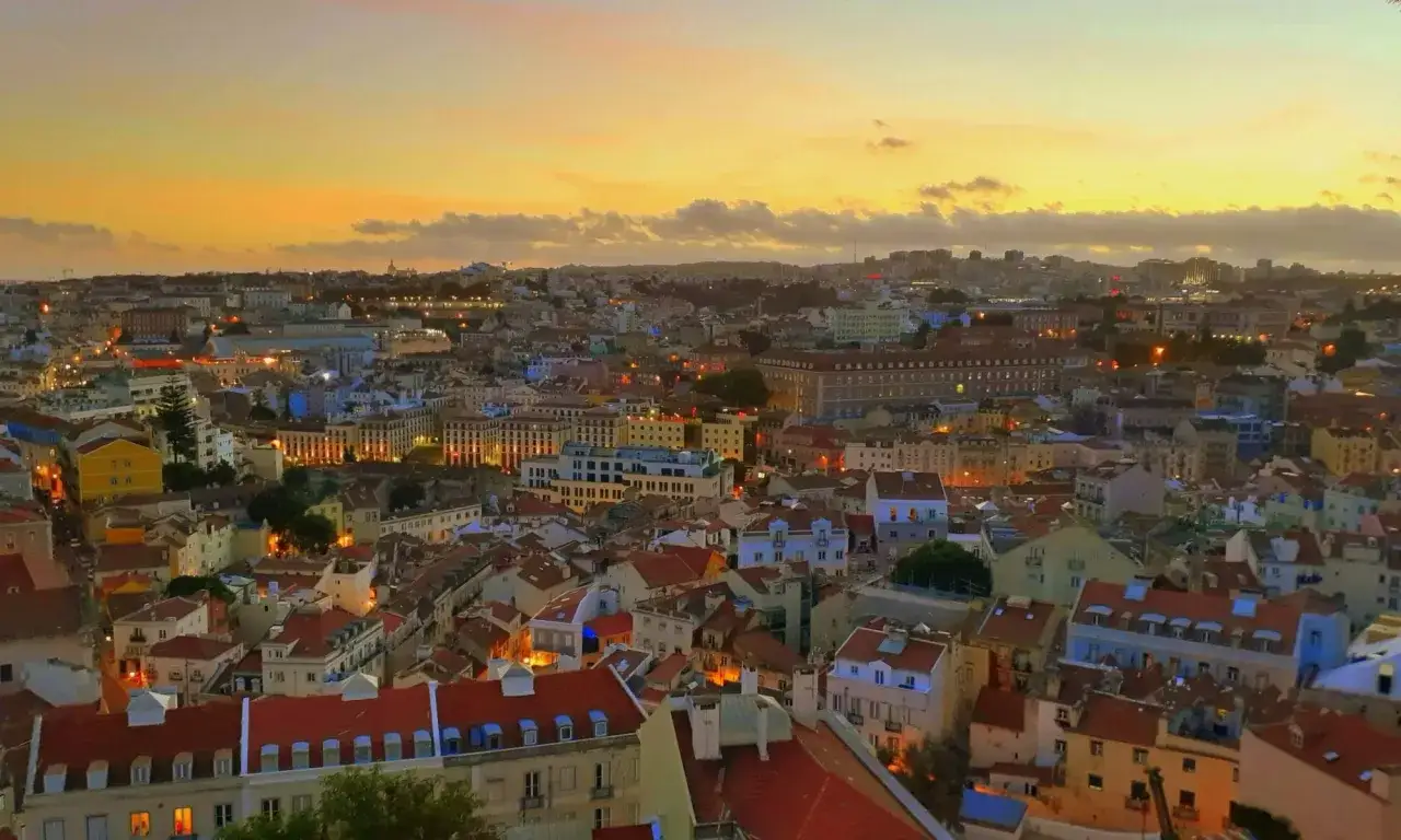 Blick vom Miradouro Lissabon bei Sonnenuntergang. Die Stadt erstrahlt in warmen Lichtern unter einem orangefarbenen Himmel.