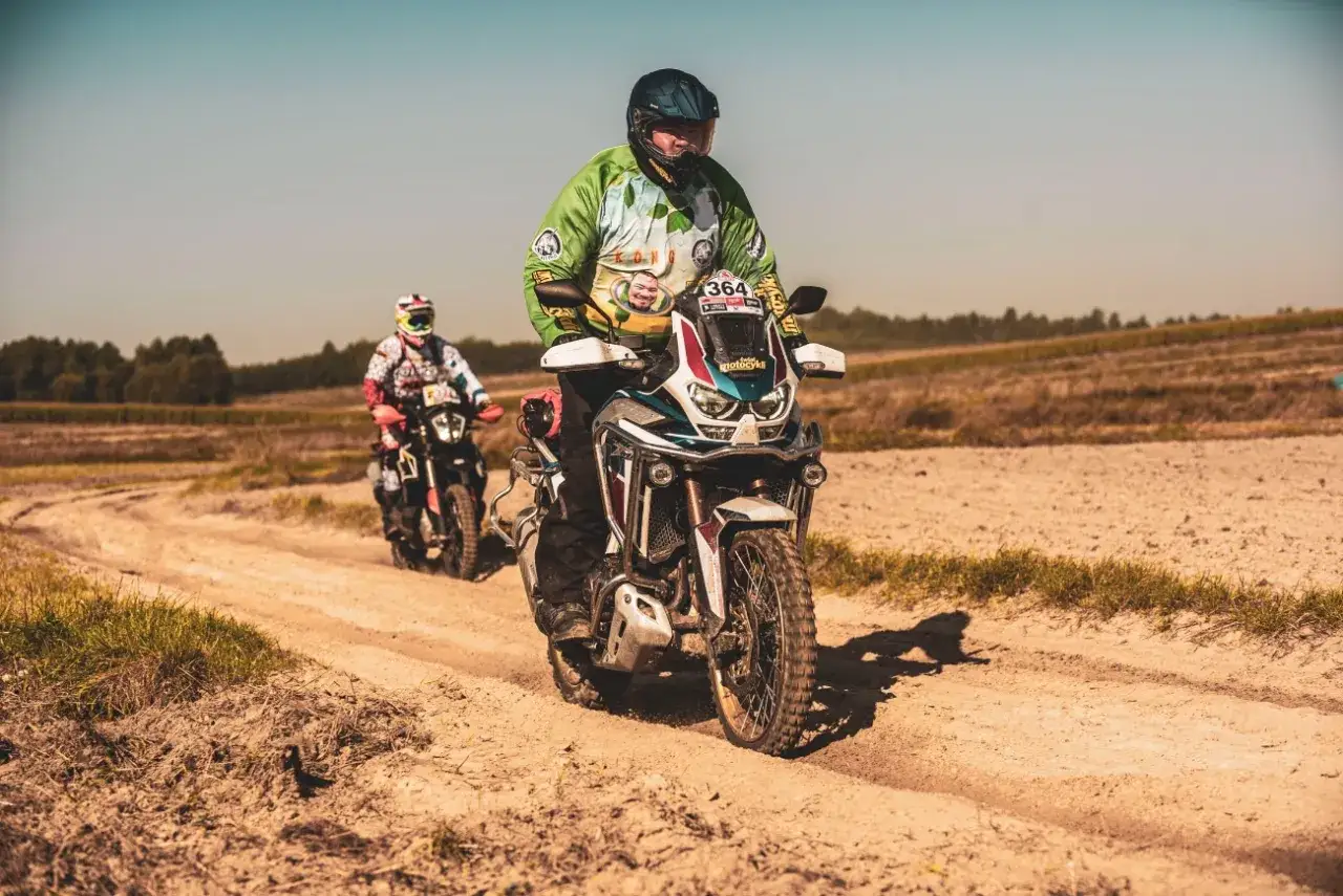 Dw&oacute;ch motocyklist&oacute;w na trasie, jeden z nich na motocyklu do lekkie enduro, pokonuje piaszczystą drogę w słoneczny dzień.