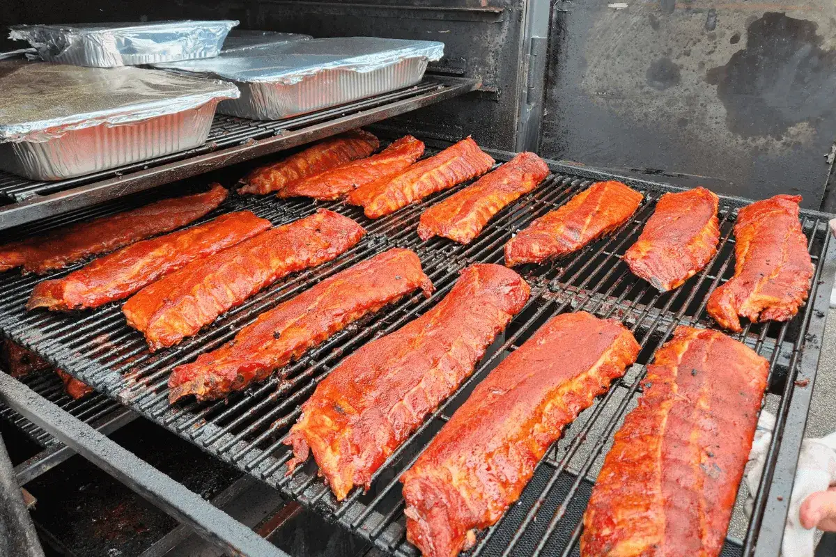 Des c&ocirc;tes lev&eacute;es de porc marin&eacute;es au barbecue sur une grille. Des plats en aluminium recouverts sont empil&eacute;s au-dessus.