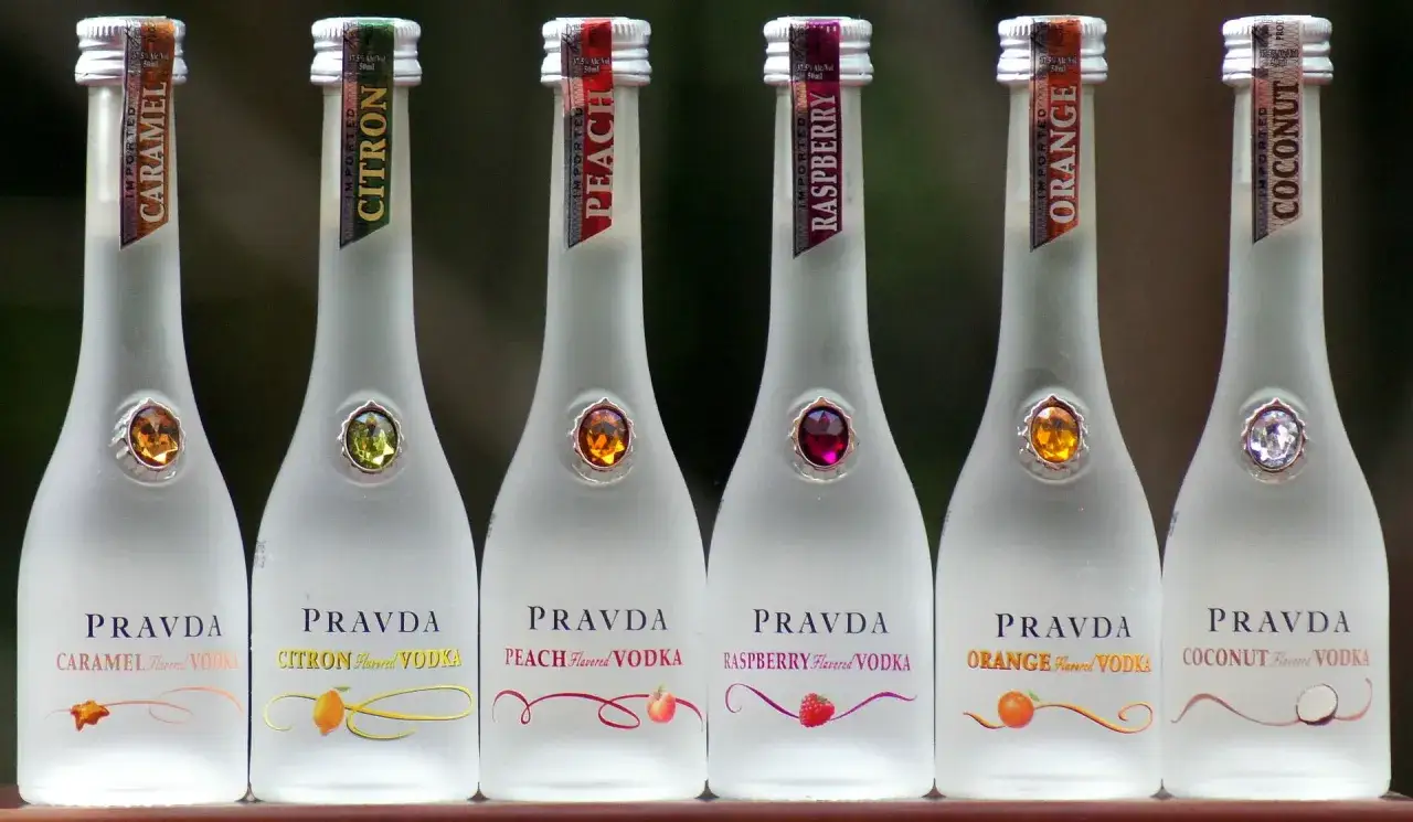 Rząd matowych butelek z różnymi **dobrymi wódkami smakowymi** Pravda: karmelową, cytronową, brzoskwiniową, malinową, pomarańczową i kokosową.