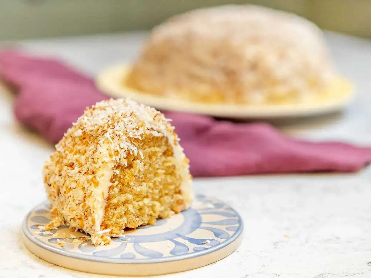 En saftig sockerkaka med kokos, täckt av rostad kokosflingor. En bit ligger på en blåvit tallrik, en hel kaka syns i bakgrunden.