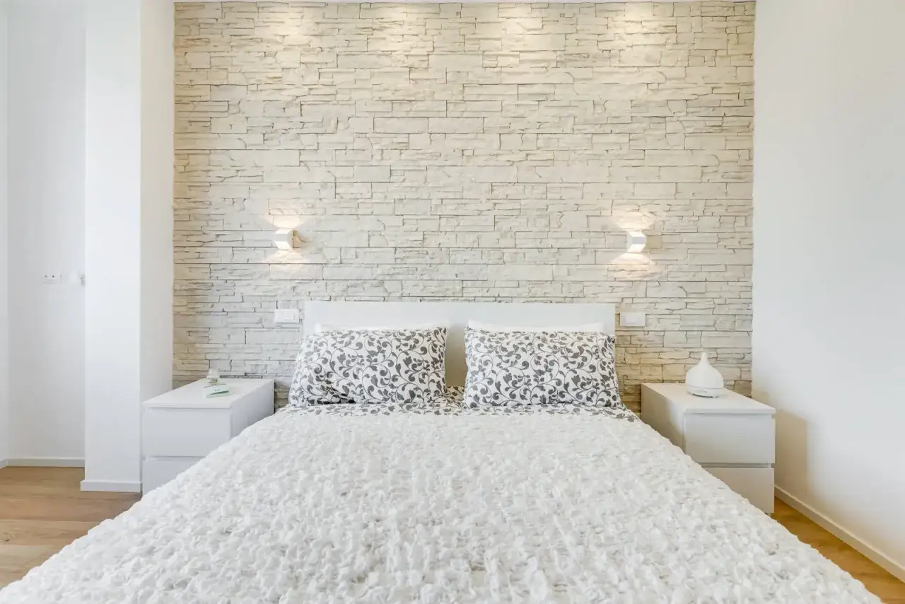 Moderna camera da letto con parete in pietra chiara, un letto bianco e due comodini. Ottime idee pareti in pietra per un tocco naturale.