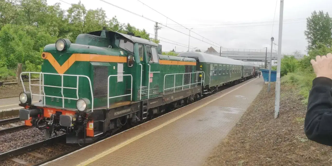 Zielona lokomotywa SU42 z żółtym pasem stoi na torach, obok niej długi skład wagonów. Pociąg czeka na pasażerów, być może wyruszy z Konina do Łodzi.