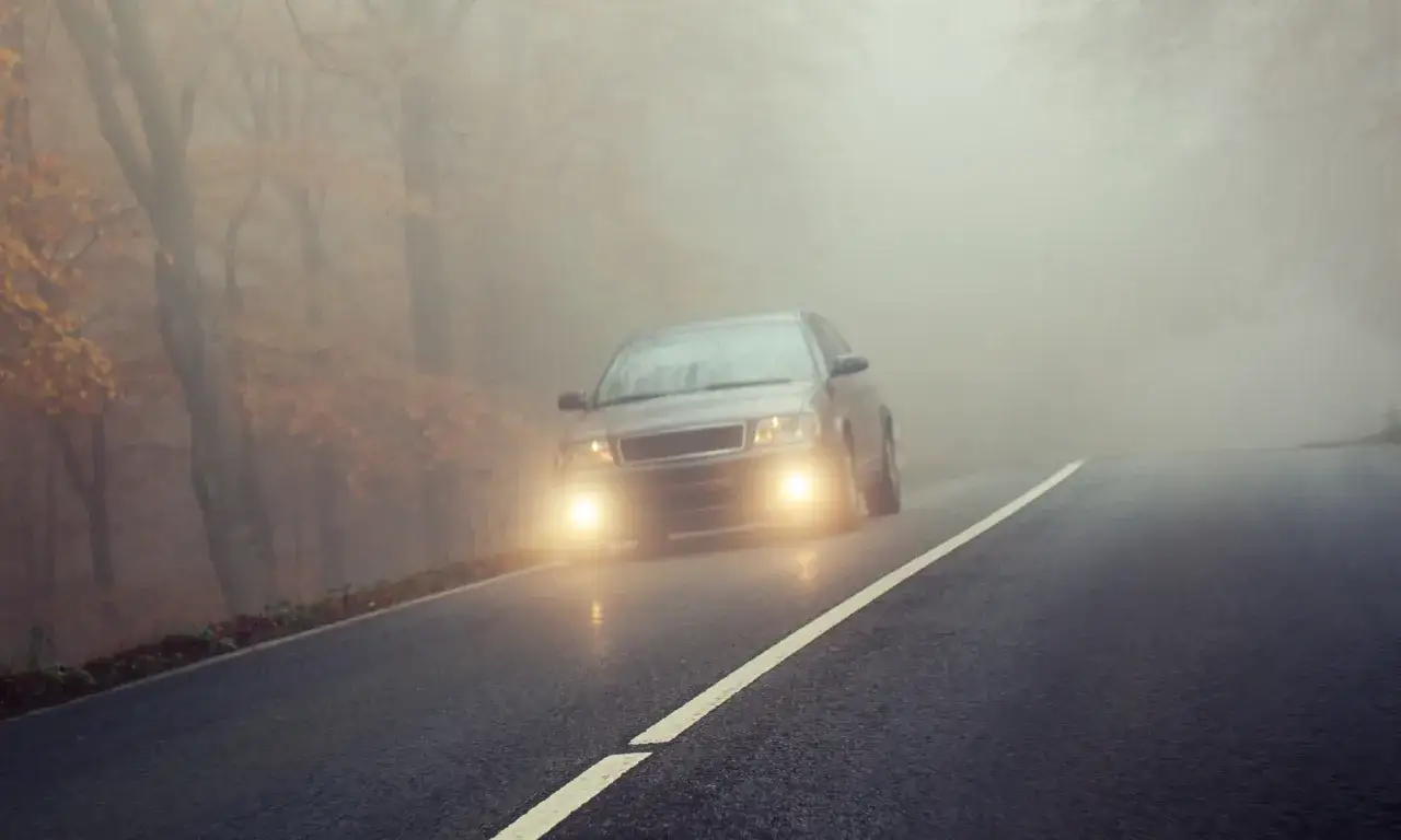 Un coche avanza por una carretera neblinosa, sus luces antiniebla iluminan el camino.