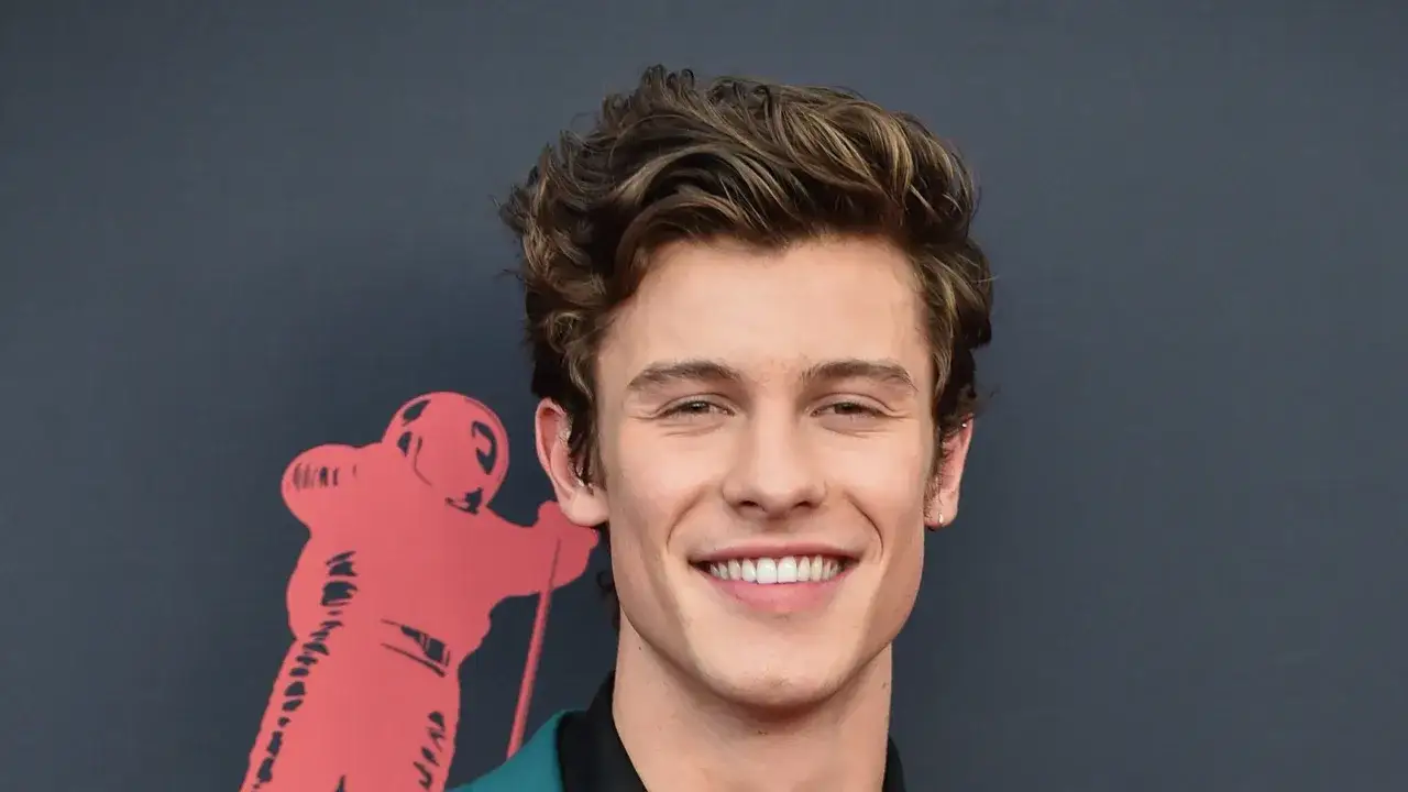 Sonriente hombre con pelo ondulado hombre, con un fondo gris y un logo rojo a la izquierda.