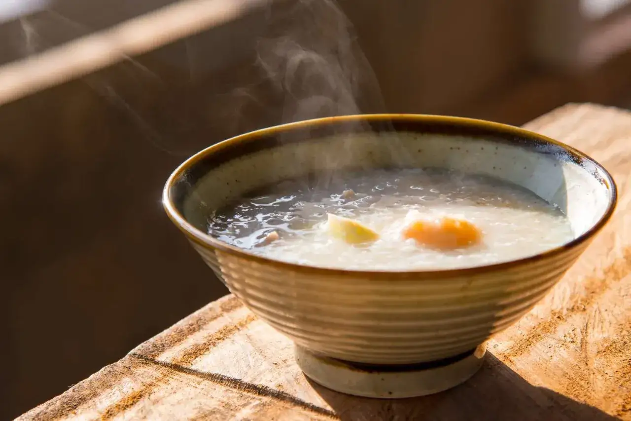 Parująca miska congee z jajkiem i kawałkami imbiru, podana na drewnianym blacie.
