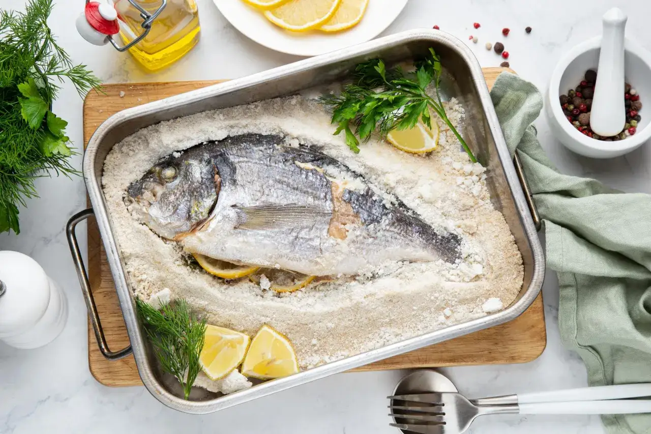 Dorada w soli, zapiekana z cytryną i ziołami, czeka na podanie. Obok oliwa, przyprawy i świeży koperek.