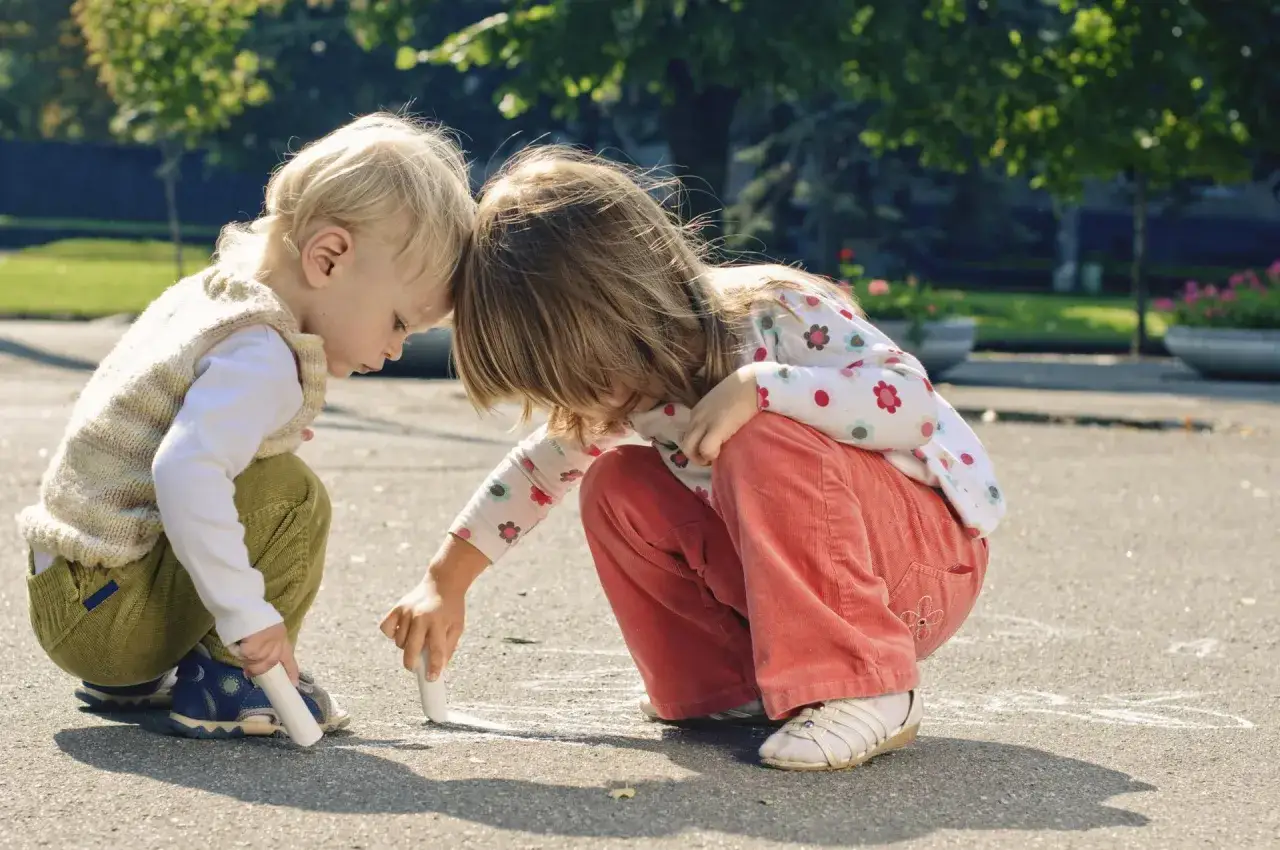 Dwie dziewczynki rysują kredą na chodniku. Ich bliskość sugeruje, że tworzą razem bezpieczną przestrzeń, jak w rodzinie zastępczej czy po adopcji.