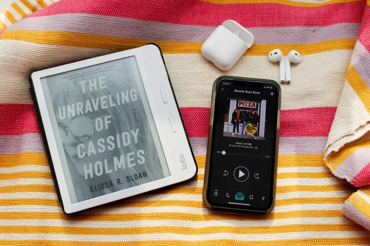 Un livre témoignage sur un Kobo, un smartphone avec un podcast et des écouteurs sans fil, le tout sur une serviette rayée.