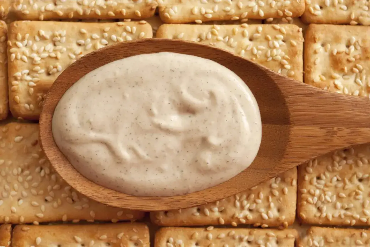 Cremosa salsa tahini que es, servida en cuchara de madera sobre galletas de sésamo.