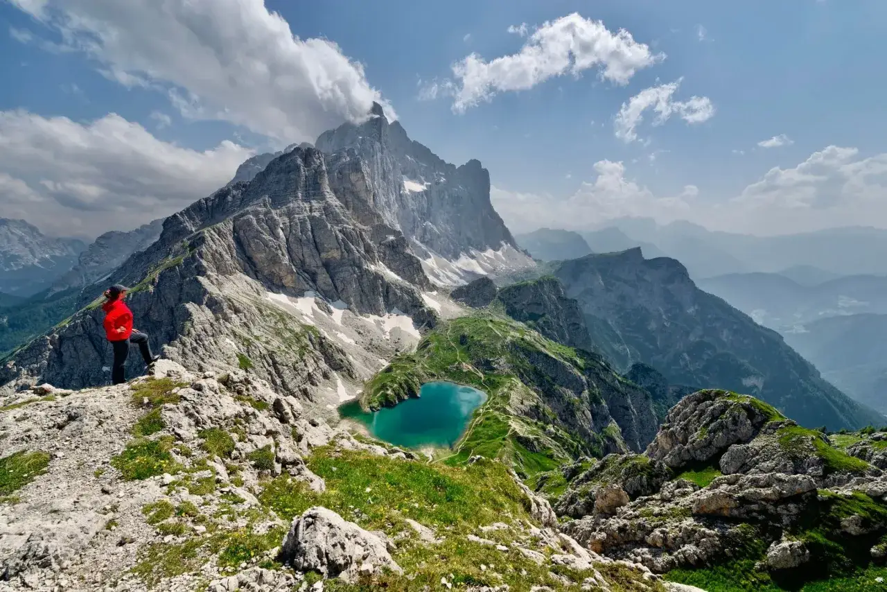 A hiker in a red jacket stands on a rocky outcrop, overlooking a stunning turquoise lake nestled in the Italian Dolomites. The majestic peaks of the Dolomites provide a dramatic backdrop to this breathtaking hiking scene.