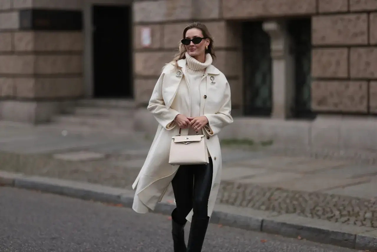 Elegancka kobieta w kremowym płaszczu, golfie i skórzanych legginsach, z beżową torebką. Stylowe skórzane legginsy stylizacje na co dzień.