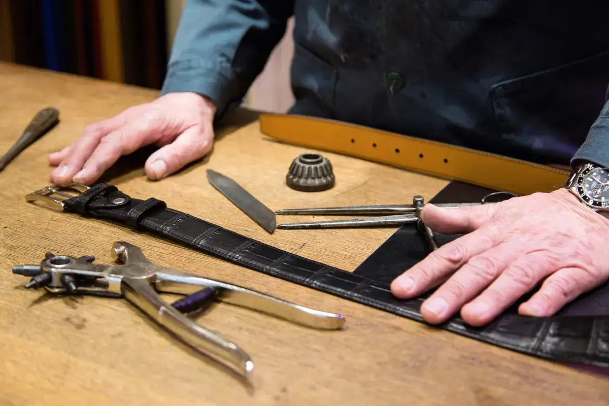 Un artisan utilise un outil cordonnier pour travailler sur une ceinture en cuir noir. Divers outils sont dispos&eacute;s sur la table en bois.