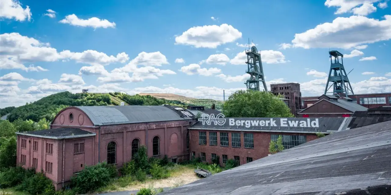 Industriearchitektur des **Steelpark Ewald** mit Fördertürmen und Backsteingebäuden unter blauem Himmel mit Wolken.