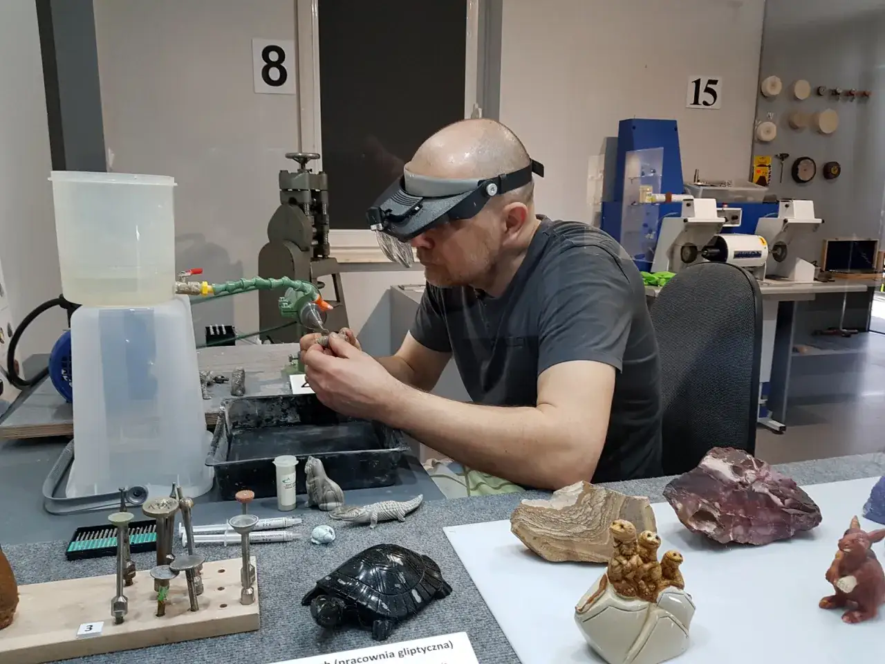 Mężczyzna w okularach ochronnych z lupą precyzyjnie zajmuje się szlifowaniem kamieni szlachetnych. Na stole widać narzędzia i gotowe wyroby.