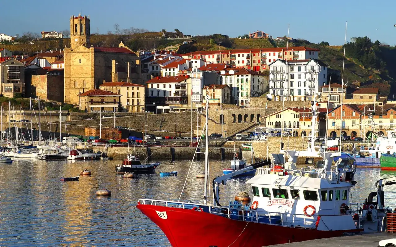 Puerto de Getaria, cerca de San Sebastián, con barcos de pesca rojos y blancos en primer plano y un pueblo pintoresco en la colina.