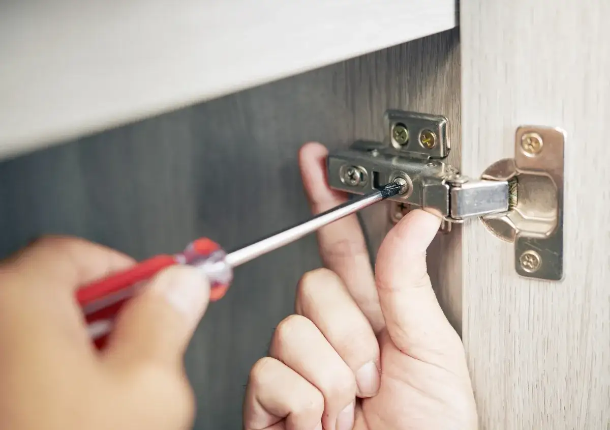A hand uses a screwdriver to remove a screw from a cabinet hinge, demonstrating how to remove stuck screws.