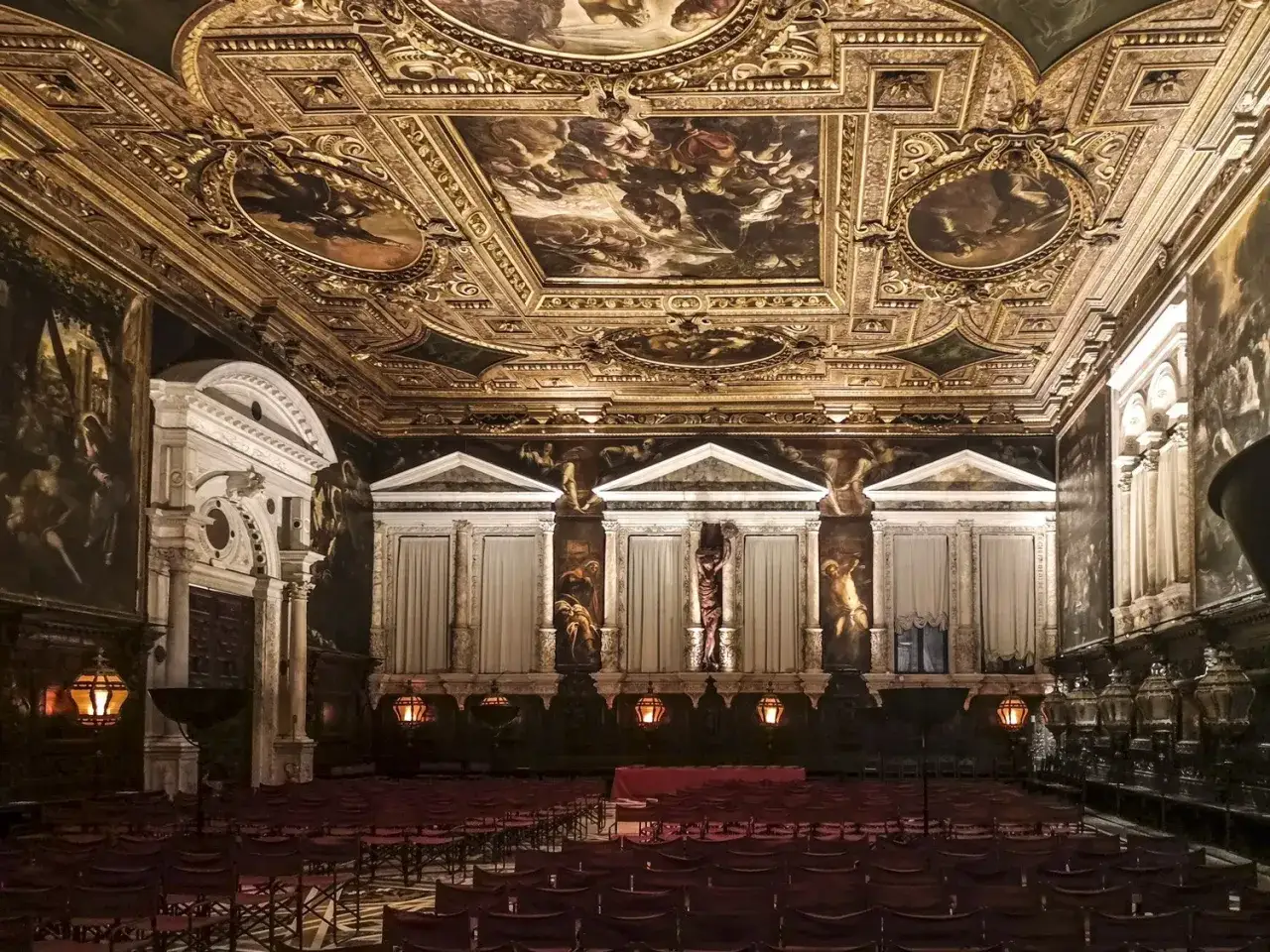 La sala capitolare della Scuola Grande di San Rocco, con soffitto dorato affrescato e file di sedie rosse.