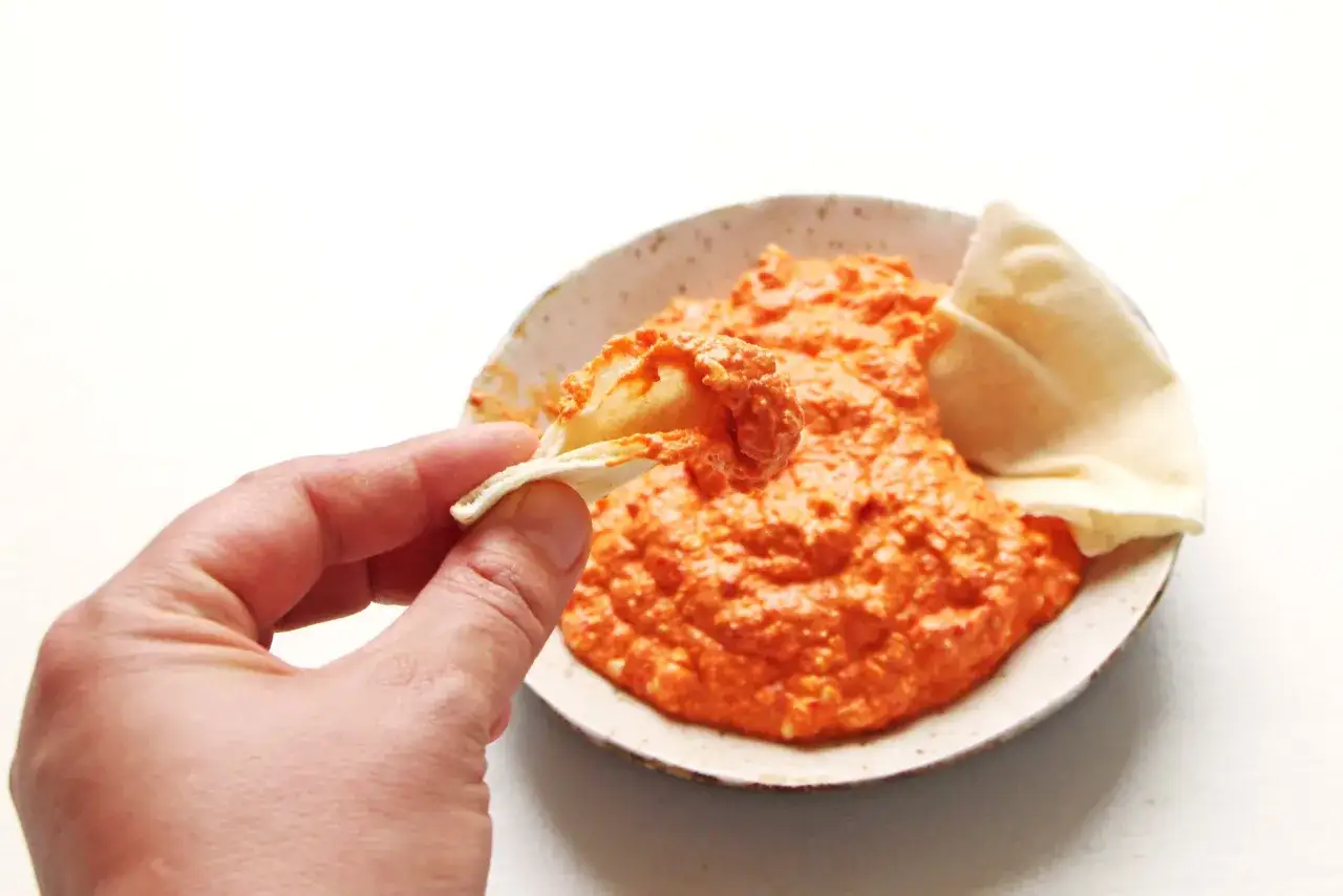 Une main trempe un morceau de pain pita dans une pur&eacute;e orang&eacute;e, probablement &agrave; base de poivron rouge.