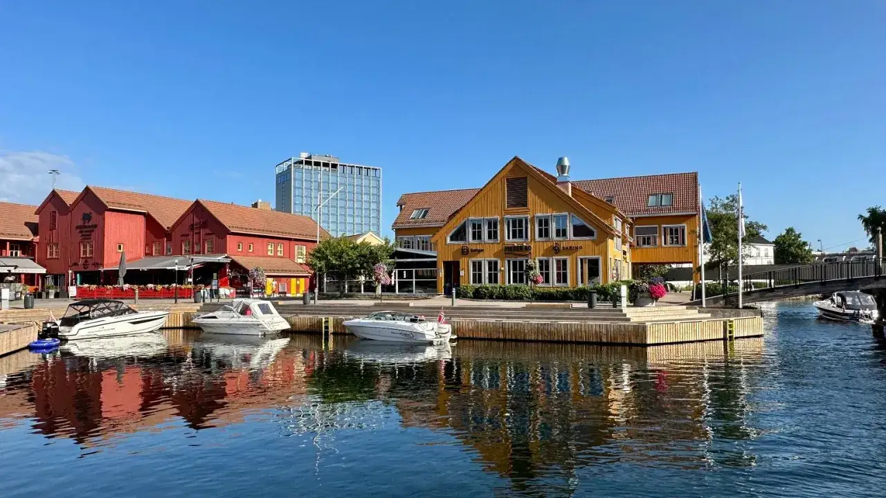 Malowniczy port w Kristiansand, Norwegia, z czerwonymi i żółtymi budynkami, łodziami na wodzie i nowoczesnym wieżowcem w tle.