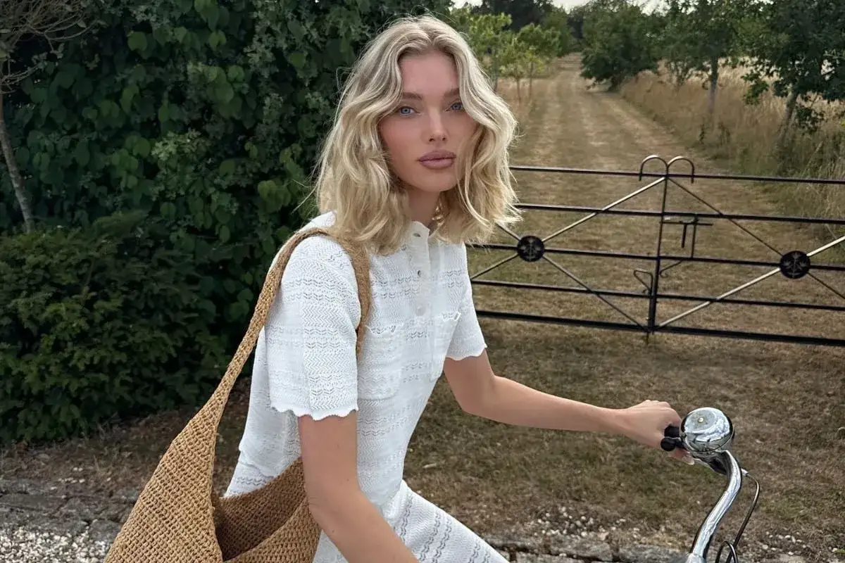 En kvinna med blont h&aring;r cyklar i ett lantligt landskap, kl&auml;dd i ett vitt linne sett och med en stor str&aring;v&auml;ska.