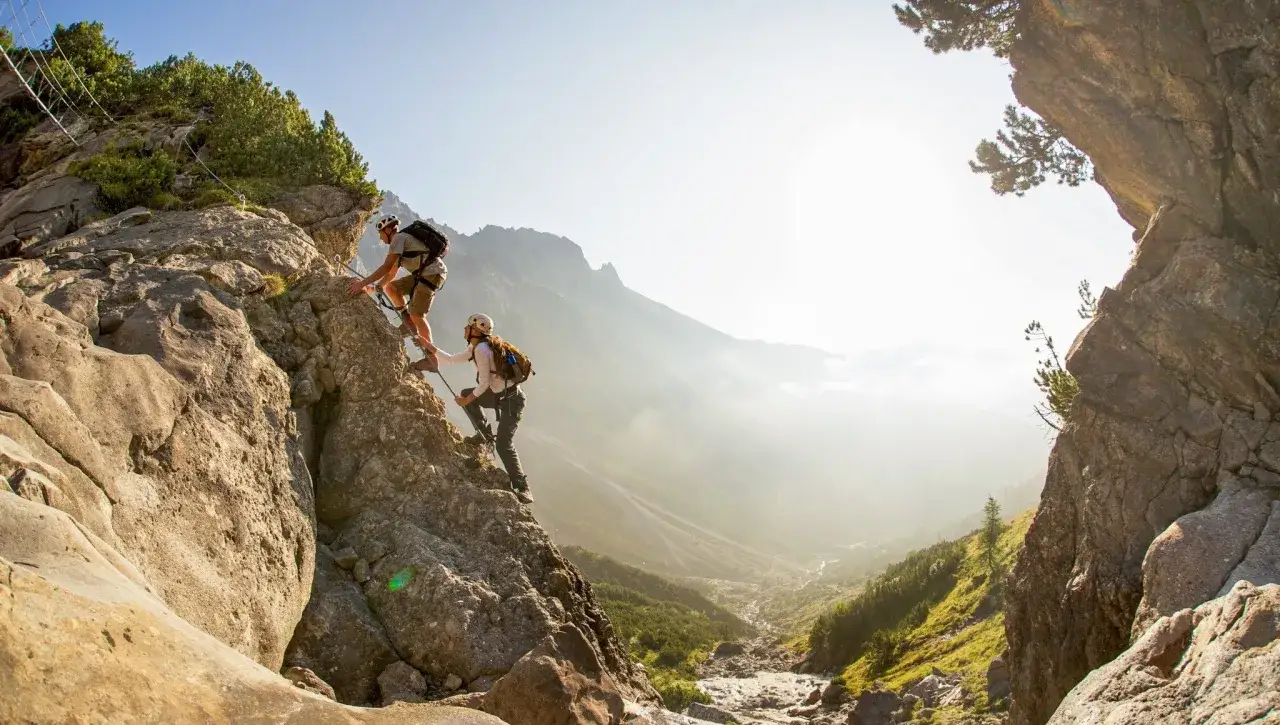 Dwóch wspinaczy pokonuje skalną ścianę na via ferrata w Austrii. Słońce rozświetla dolinę poniżej.