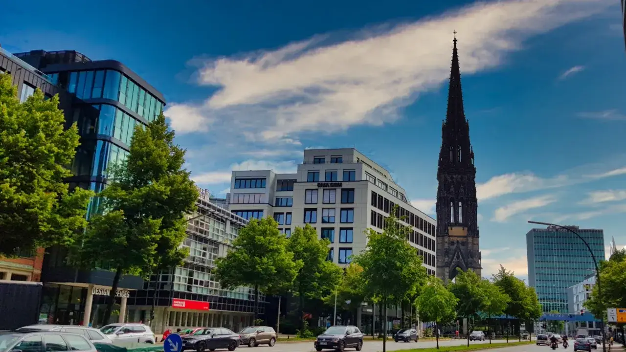 Das Mahnmal St. Nikolai ragt &uuml;ber moderne Geb&auml;ude und B&auml;ume in Hamburg. Autos fahren auf der Stra&szlig;e.