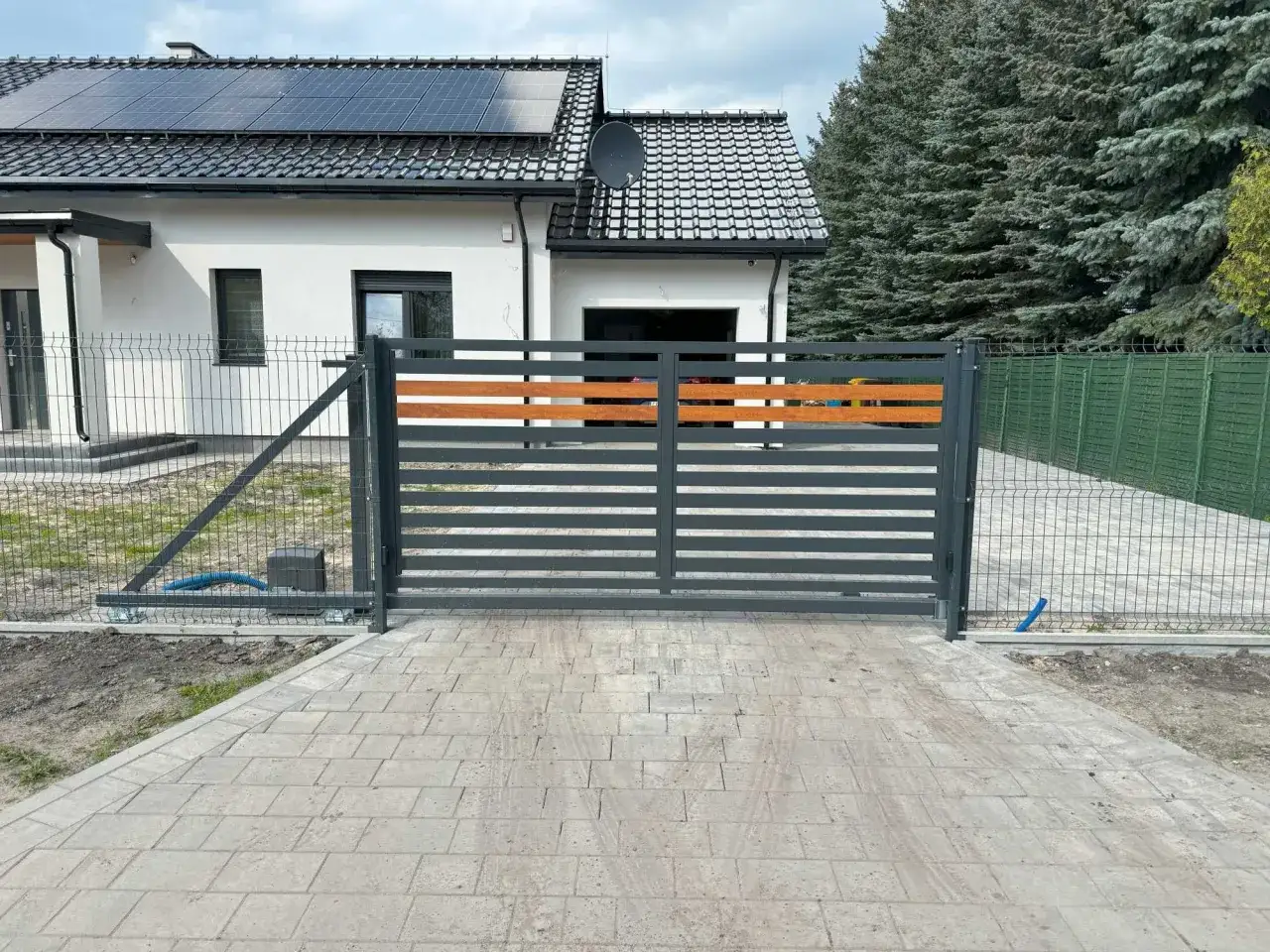 Nowoczesna, szara brama do ogrodzenia panelowego z drewnianymi wstawkami, prowadząca do domu z panelami słonecznymi.