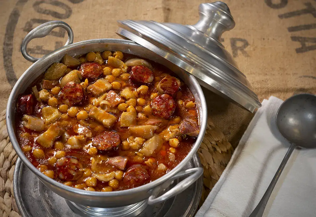Un plato humeante de callos a la gallega, con garbanzos, chorizo y trozos de carne en una cazuela met&aacute;lica.