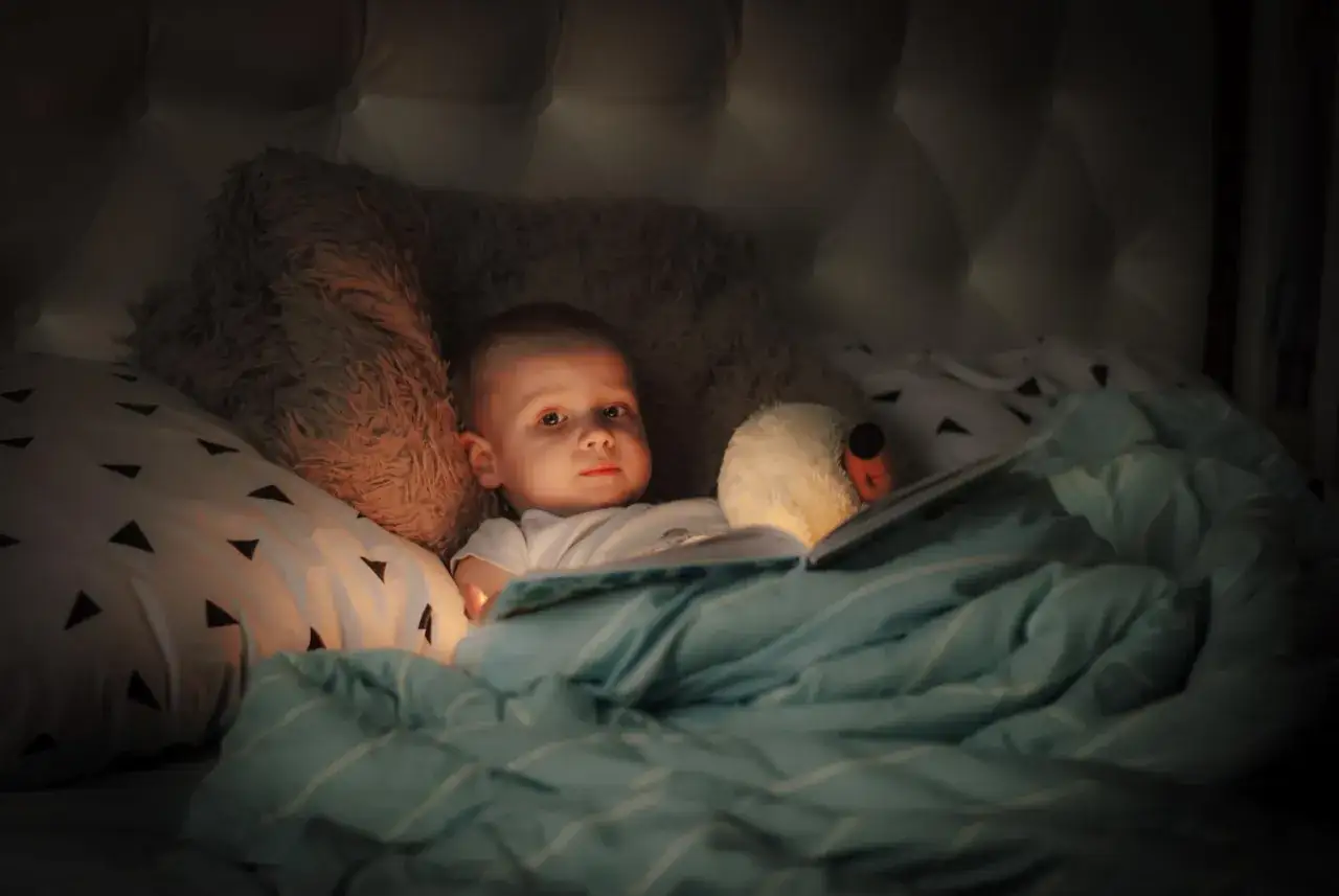 Un bimbo curioso, tra coperte e peluche, legge un libro illuminato, uno dei tanti bambini che si svegliano di notte.