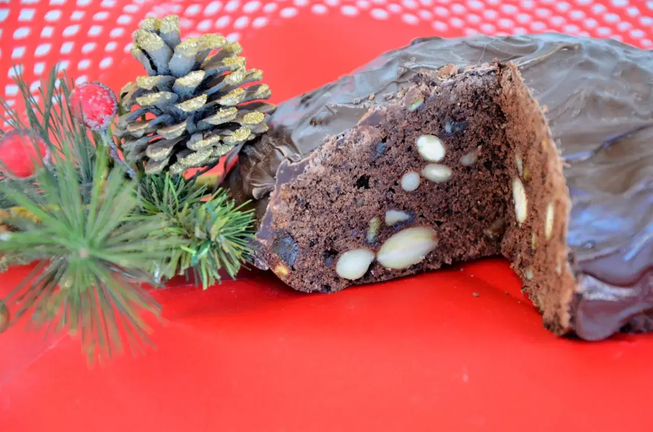 Fetta di pan pepato al cioccolato con mandorle, decorato con pigne e rami di pino, su sfondo rosso.