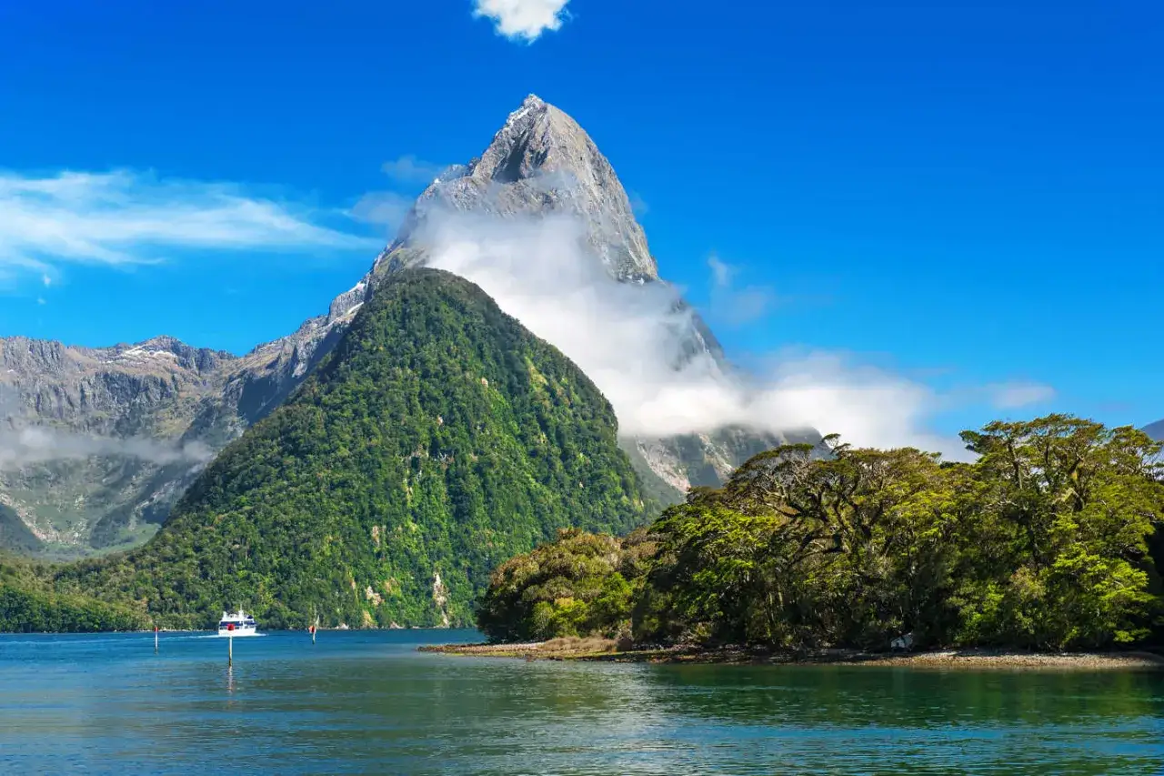 Ein Boot fährt auf einem ruhigen See, umgeben von üppig grünen Bergen. Im Hintergrund ragt ein majestätischer Gipfel empor, der als steilster Berg der Welt gilt, umhüllt von Wolken.