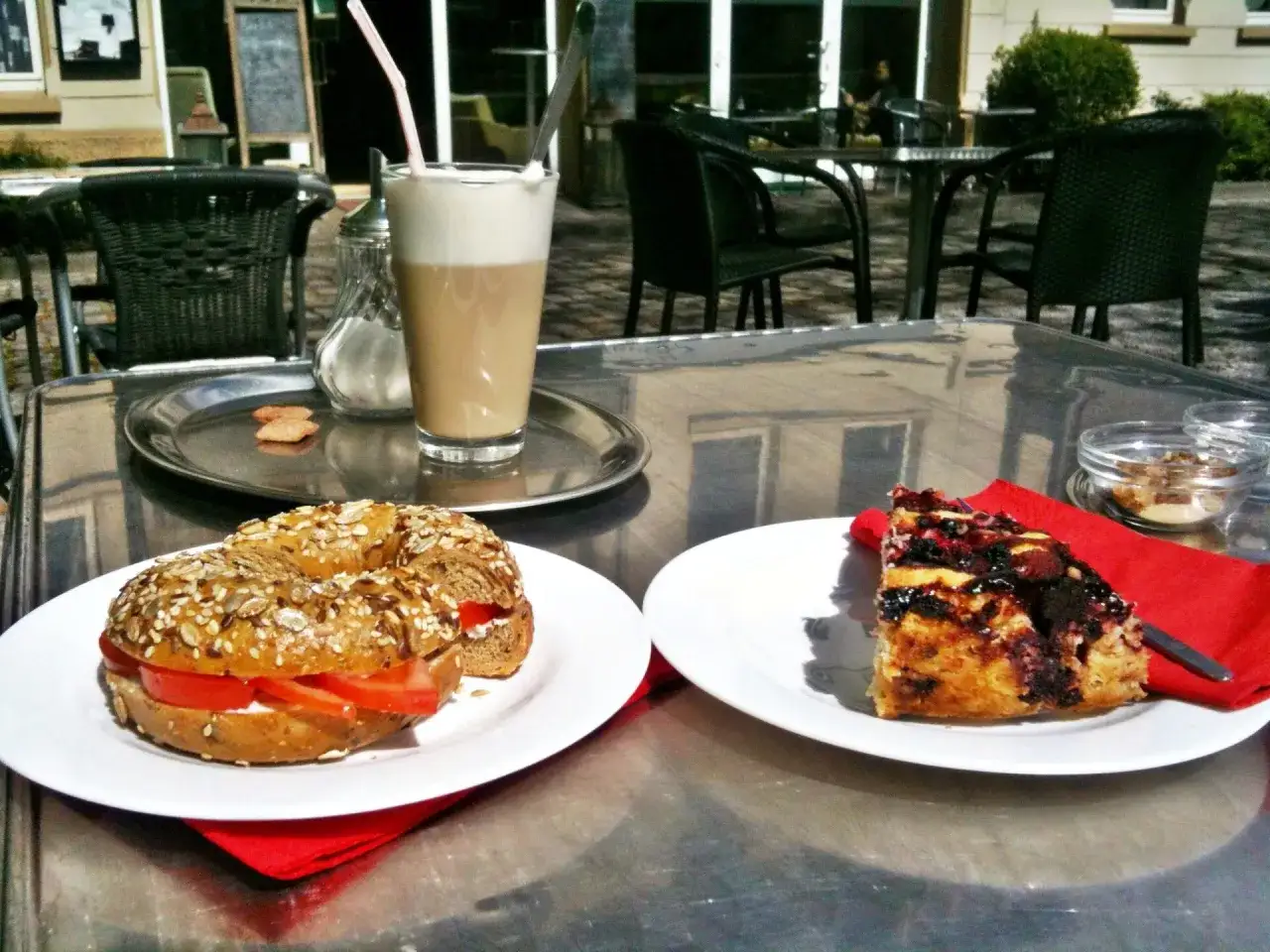 Ein veganes Cafe-Erlebnis: Bagel mit Tomate und Frischk&auml;se, ein St&uuml;ck Kuchen und ein Latte Macchiato auf einem Tisch im Freien.