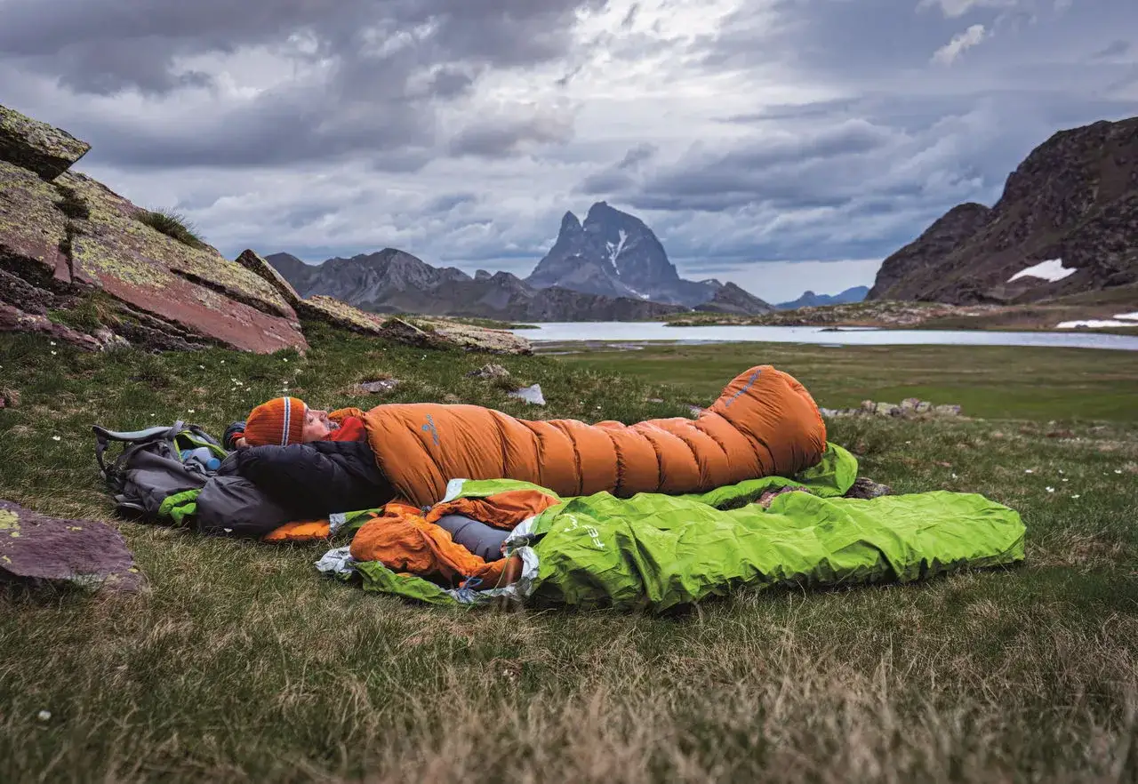 Persona durmiendo en un saco de dormir naranja, disfrutando de un vivac acampada junto a un lago y monta&ntilde;as.