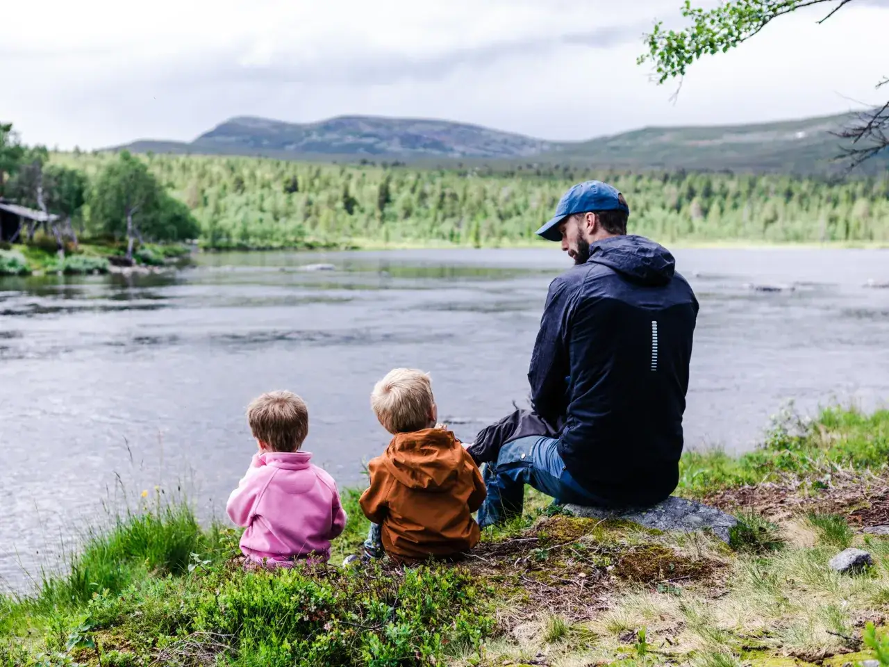 En pappa och tv&aring; barn njuter av en paus under sin vandring vid en sj&ouml;.