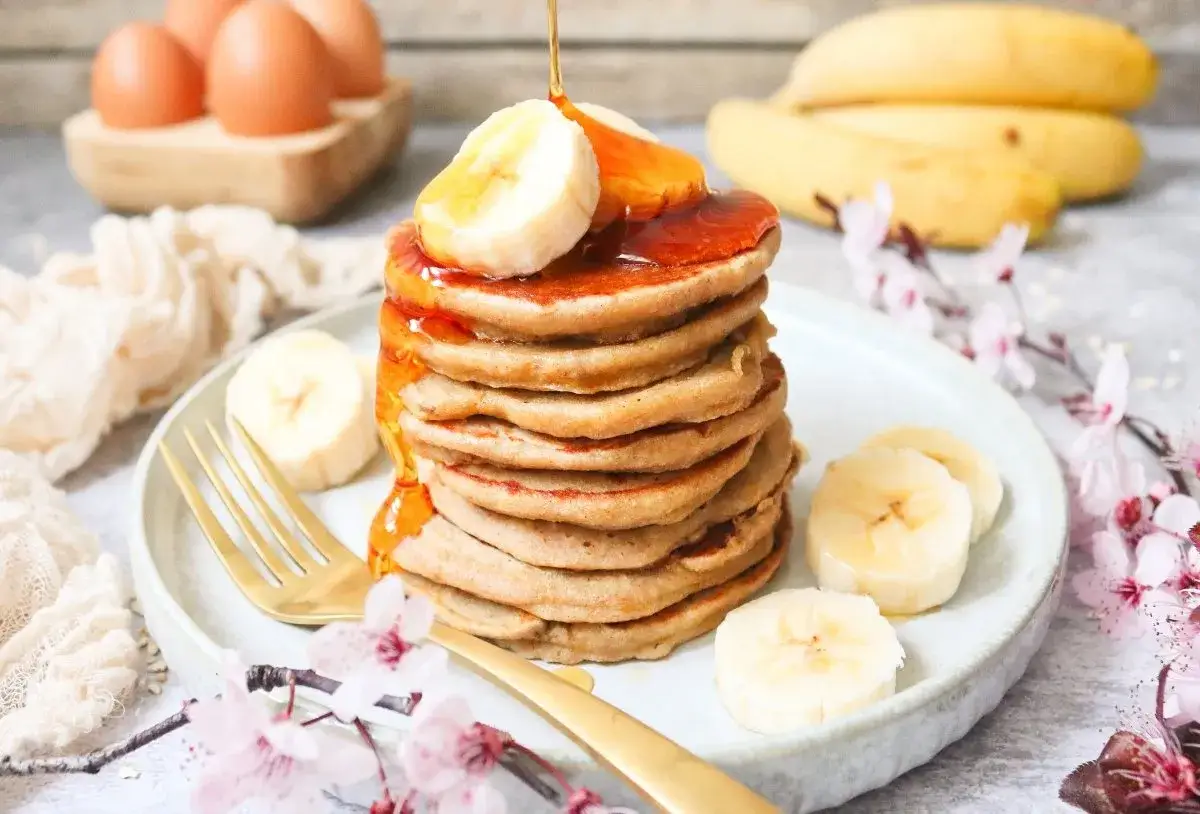 Deliciosas tortitas de avena apiladas, cubiertas con rodajas de plátano y sirope dorado. Huevos y plátanos al fondo.