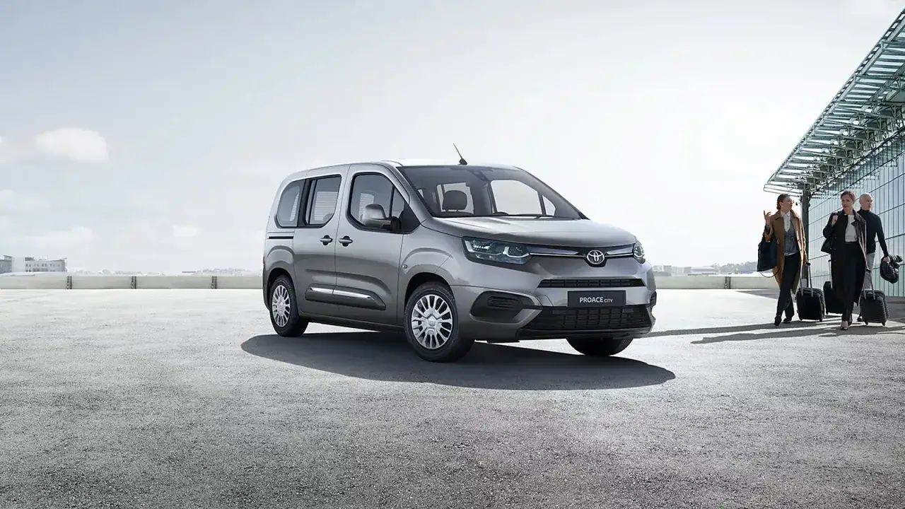 Srebrna toyota 7 osobowa czeka na pasażerów przy lotnisku. Rodzina z bagażami zmierza do samochodu.