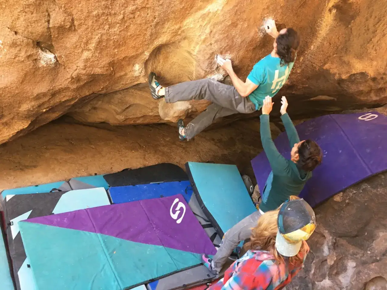 Dwóch wspinaczy na skale, jeden w powietrzu, drugi asekuruje. Wokół kolorowe materace do boulderingu.
