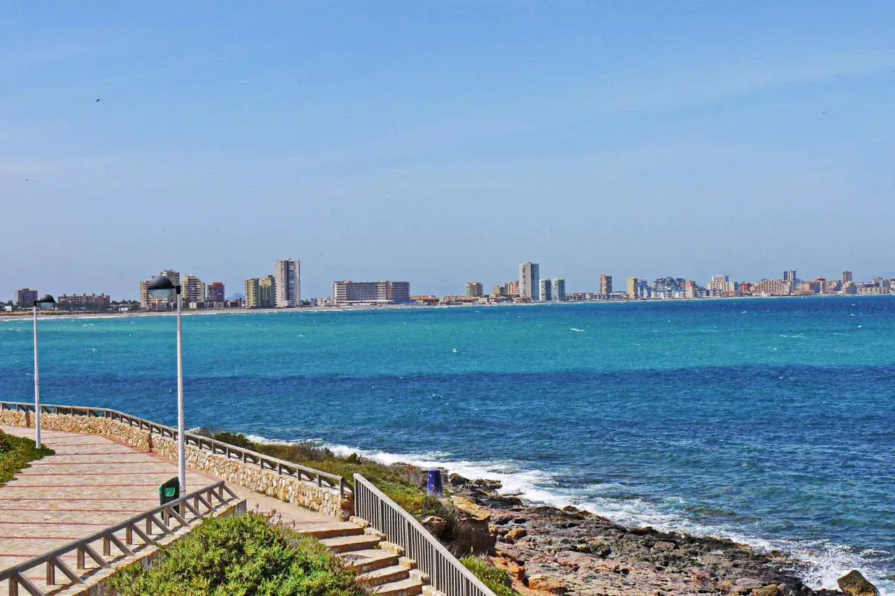 Blick auf die K&uuml;stenlinie des Mar Menor mit einer Promenade und einer Stadt im Hintergrund.