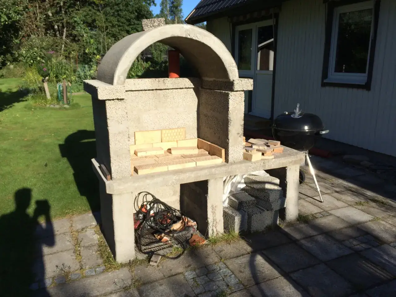 P&aring;g&aring;r bygga grill av tegelstenar. En del av stommen &auml;r klar, med tegel i eldstaden. En klotgrill st&aring;r bredvid.