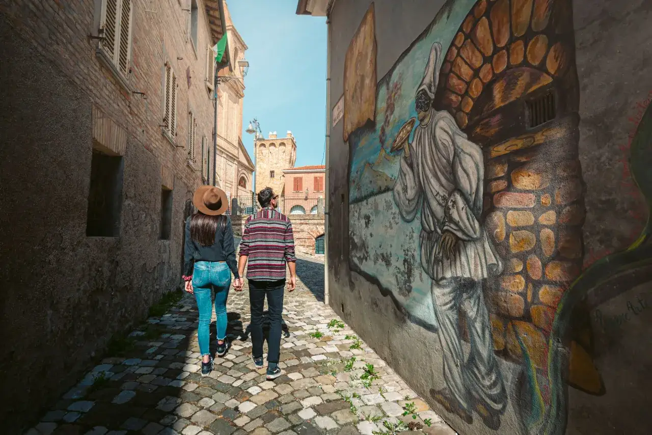 Coppia passeggia in un vicolo di Saludecio, ammirando un murale. Scopri cosa vedere in questo borgo incantevole.