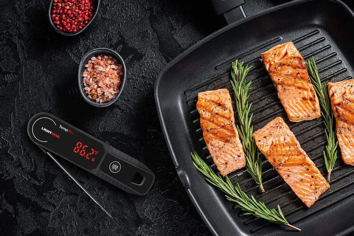 Grillad lax med rosmarin i en grillpanna, bredvid en digital termometer som visar lax innertemperatur. Små skålar med pepparkorn och flingsalt.