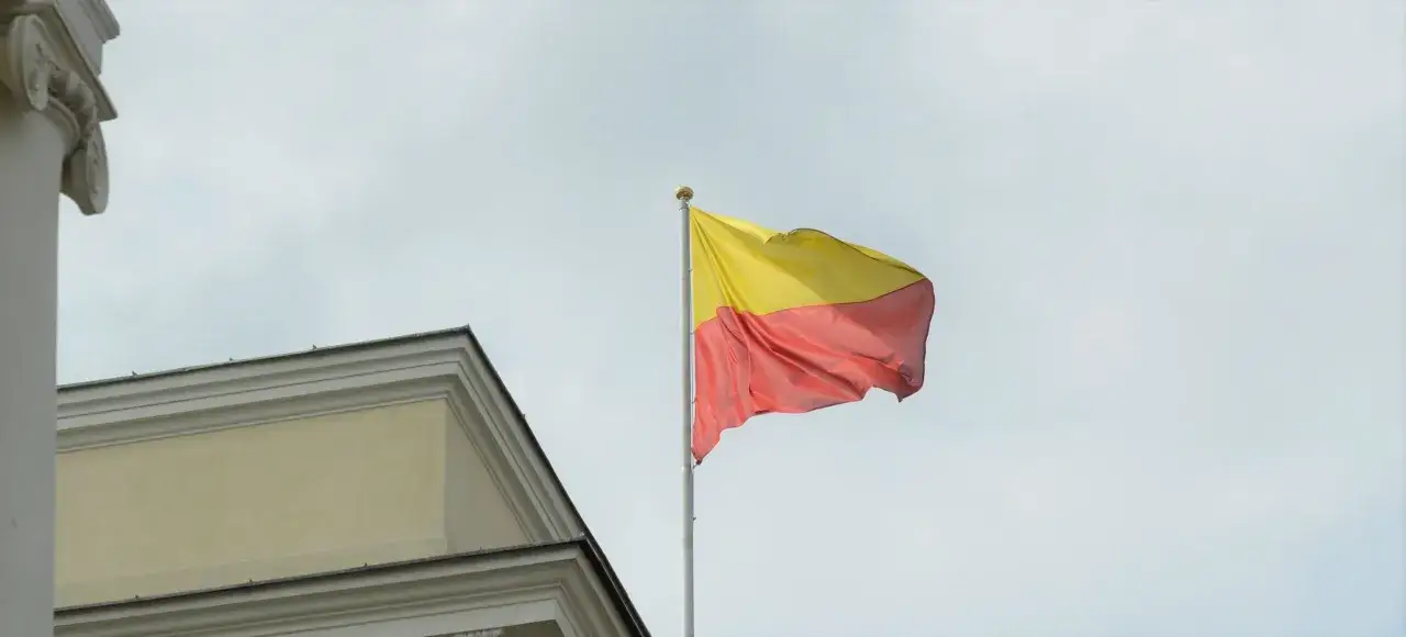 Flaga Warszawy powiewa na maszcie obok zabytkowej kamienicy.