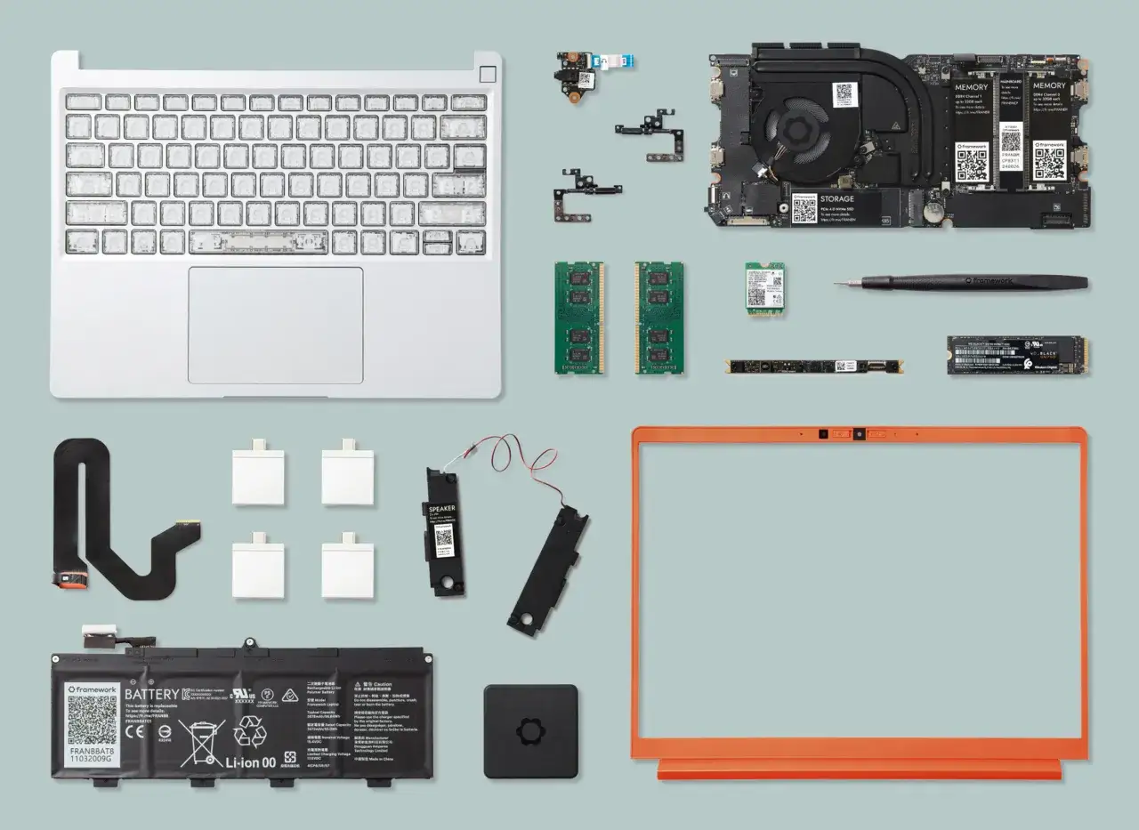 Composants d'un **framework computer** : carte mère, RAM, SSD, batterie, clavier, écran, haut-parleurs, et câbles.